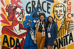 Two young women standing in front of art of Grace Hopper, Ada Lovelace, and Anita